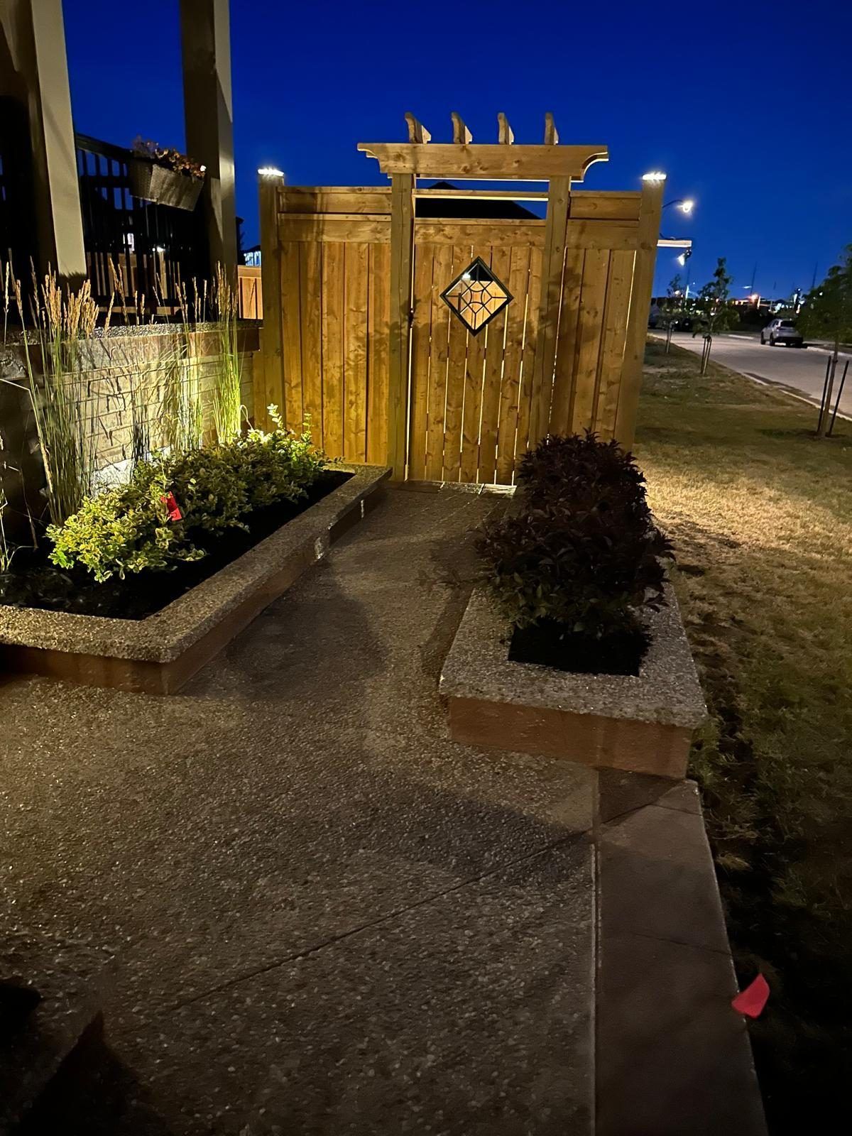 Cedar fence gate with pergola top and lit planting beds at dusk